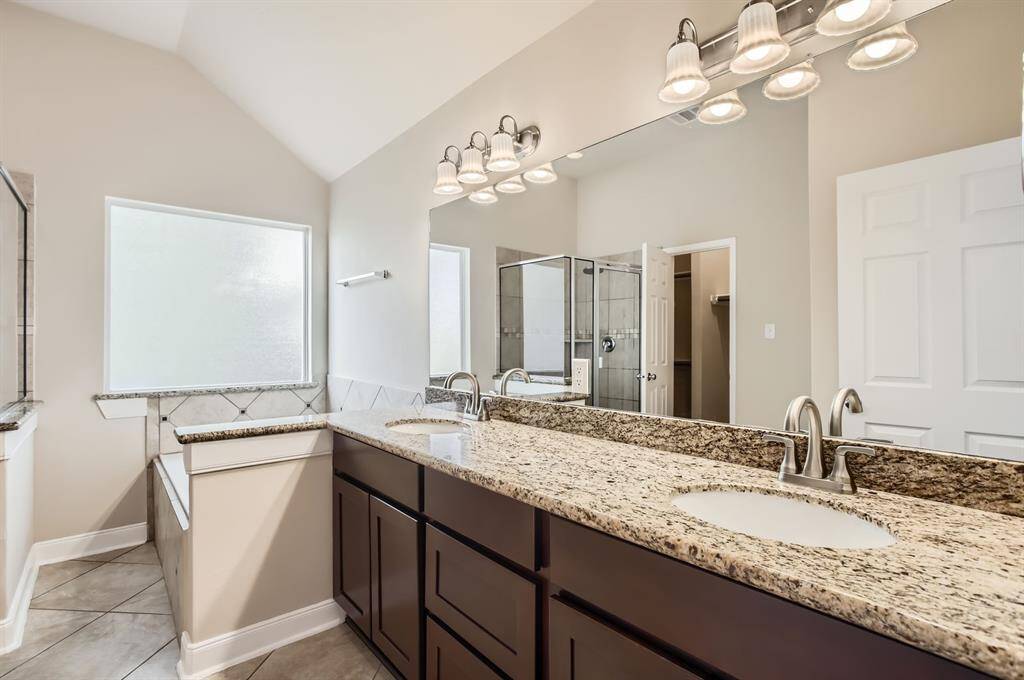 This primary bathroom features a spacious double vanity with granite countertops, modern fixtures, and ample lighting. It includes a large mirror, a glass-enclosed shower, and a separate bathtub, all set against neutral-toned tiles and walls for a clean, elegant look.