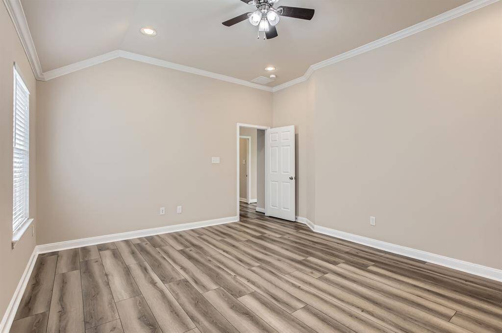 This bedroom features modern wood-look flooring, neutral walls, and crown molding. It has recessed lighting, a ceiling fan, and ample natural light from a large window. A white door leads to an adjacent space, adding to its openness.