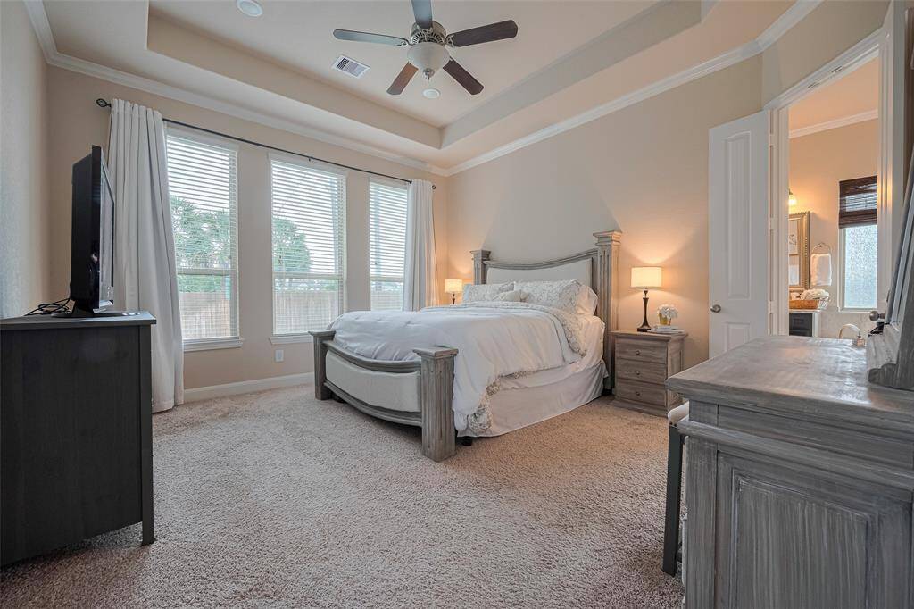 The expansive primary suite features plush carpeting, a refined tray ceiling, and multiple tall windows that bring in daylight. A ceiling fan adds an extra layer of comfort, while the attached bath enhances the sense of a personal sanctuary.