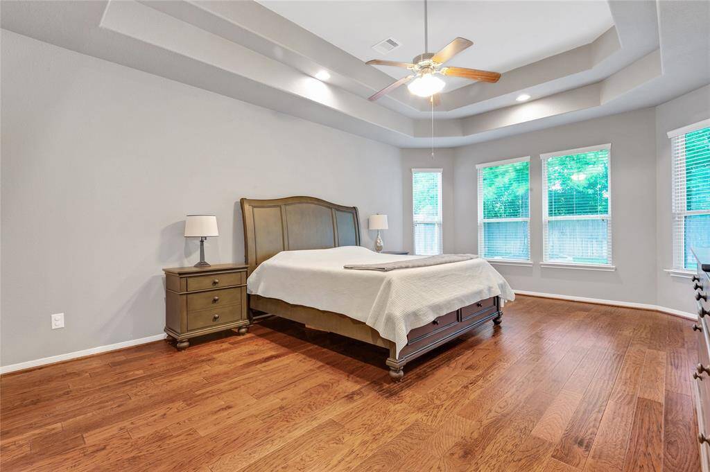 Elegant and oversized primary bedroom with the same lovely wood floors, tray ceiling, and windows overlooking the serene park like backyard.