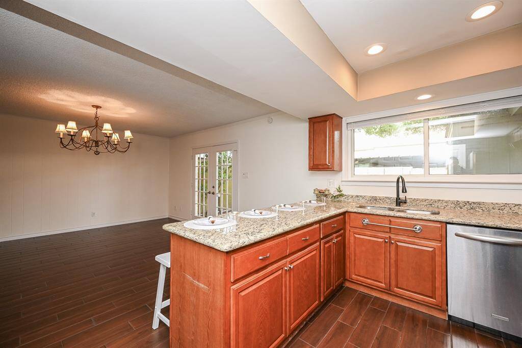This photo features a spacious open-concept kitchen and dining area. The kitchen has granite countertops, wooden cabinets, and stainless steel appliances. Large windows provide natural light, and a modern chandelier adds elegance to the dining space. French doors lead outside, enhancing the room's appeal.