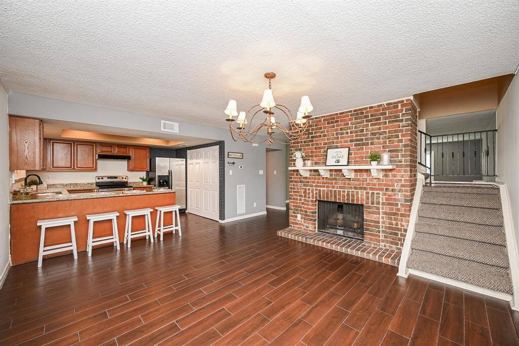 The photo showcases a cozy open-concept living area with a warm, inviting atmosphere. It features a brick fireplace, hardwood floors, and a modern kitchen with granite countertops and wooden cabinetry. The space is enhanced by a stylish chandelier and an accessible staircase, creating a perfect blend of comfort and functionality.