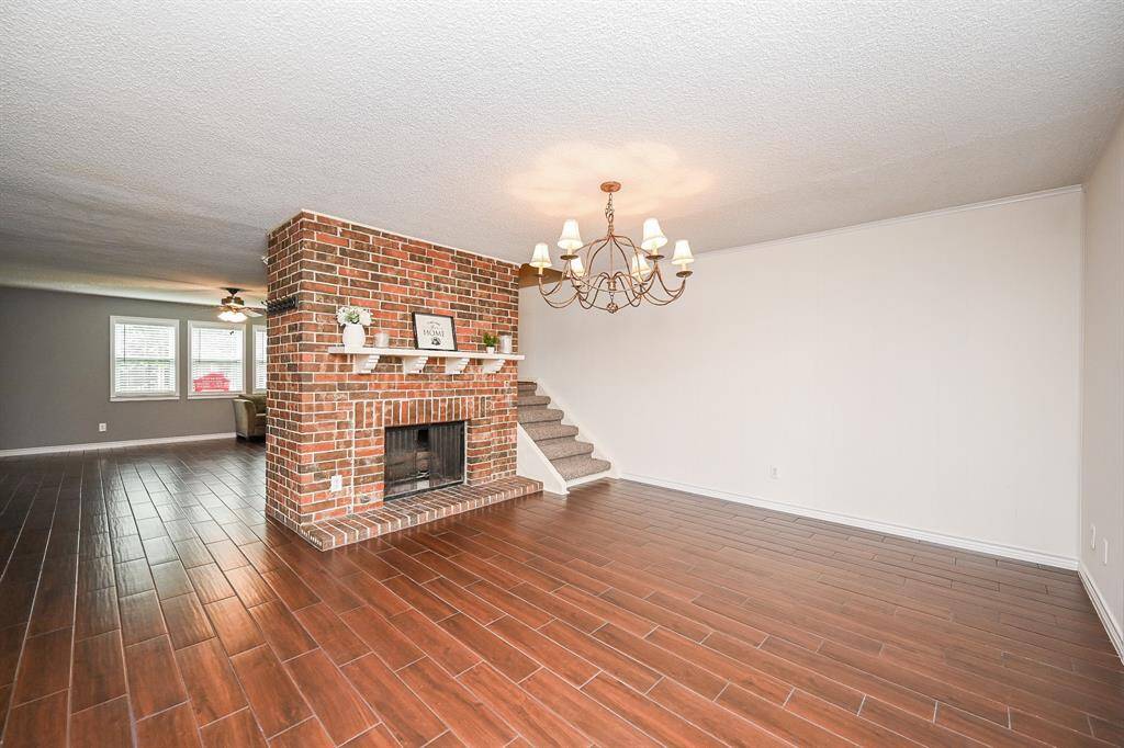 This spacious living area features a brick fireplace as a focal point, an elegant chandelier, and wood-like tile flooring. The open layout leads to the dining area, with stairs providing easy access to the upper level.