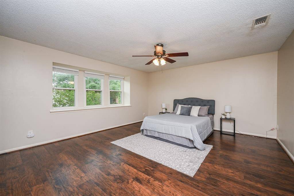 This spacious bedroom features rich hardwood floors, a large window offering plenty of natural light, and a ceiling fan for comfort. The neutral walls provide a versatile backdrop, perfect for personalizing your space.