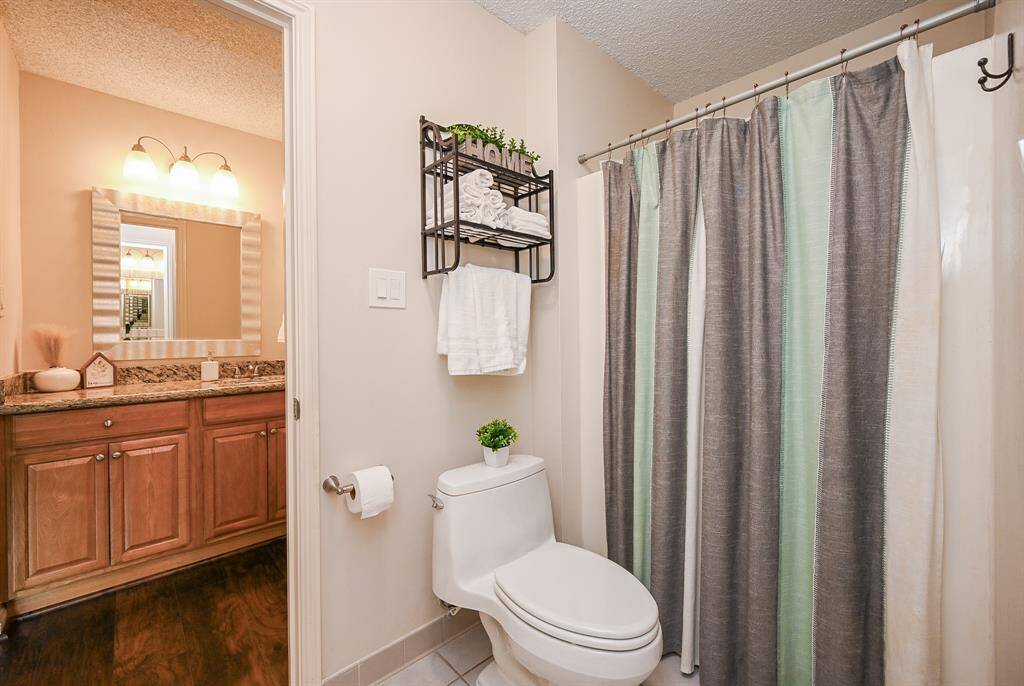 This bathroom features a modern design with a neutral color palette. It includes a granite-countered vanity with wooden cabinets, a large mirror, and a three-light fixture. The shower is enclosed with a striped curtain, and there's a wall-mounted shelf for towels above the toilet.