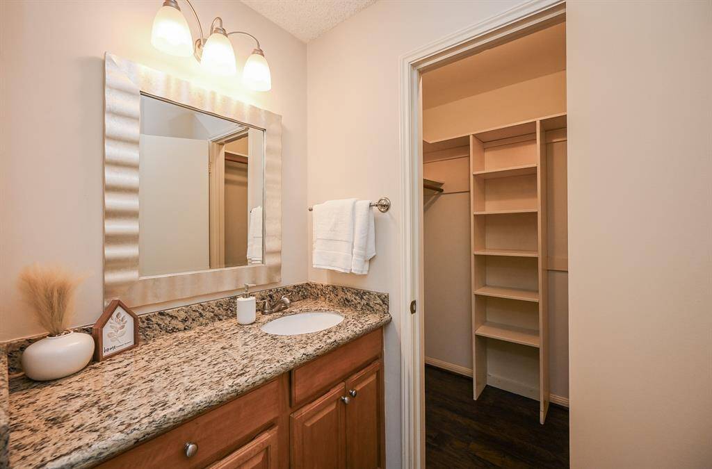 This photo shows a well-lit bathroom with a granite countertop, under-mount sink, and a large framed mirror. It opens into a spacious walk-in closet with built-in shelving.