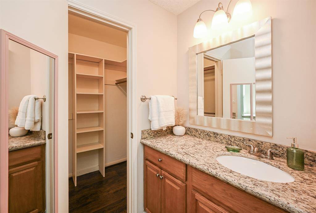 This photo showcases a bathroom with a granite countertop, a stylish mirror, and ample lighting. It includes wooden cabinetry and an adjacent walk-in closet with built-in shelving, offering plenty of storage space.