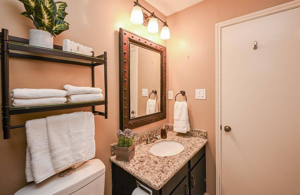 Cozy bathroom with warm tones, featuring a granite countertop, framed mirror, and modern light fixture. Includes a wall shelf for storage and decor, adding functionality and style.
