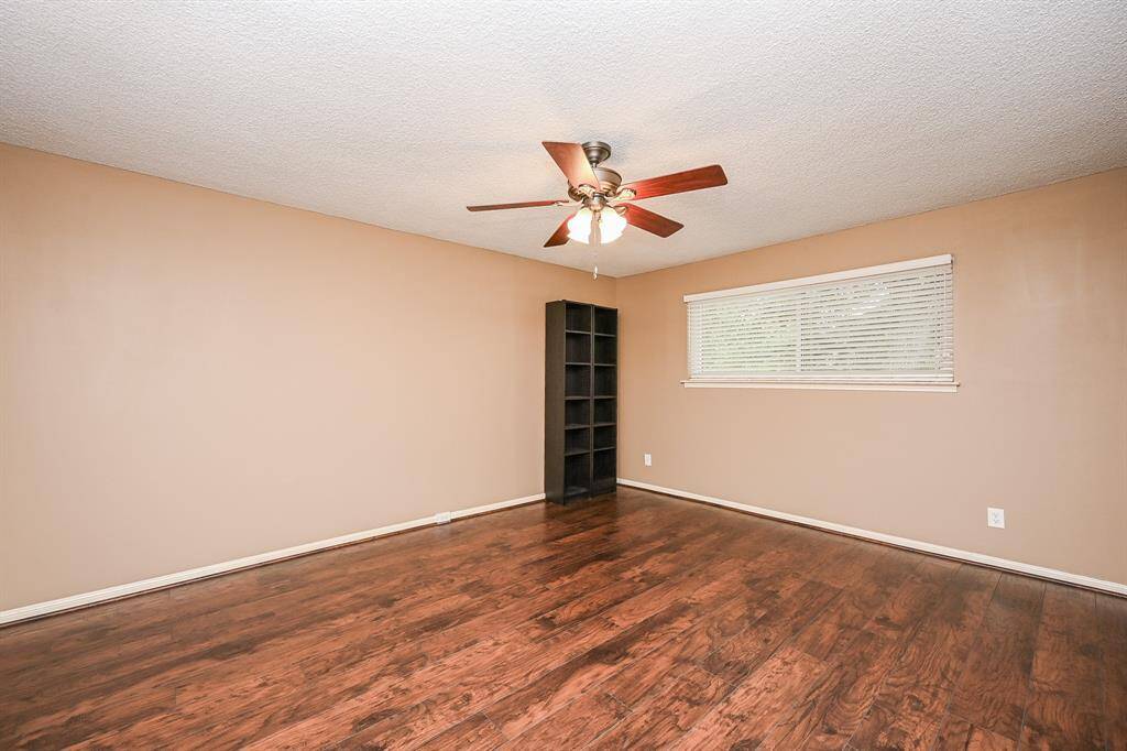 This room features warm-toned walls and rich hardwood floors. It includes a ceiling fan with lights and a large window with blinds, providing ample natural light. A built-in shelving unit is available for storage or display.