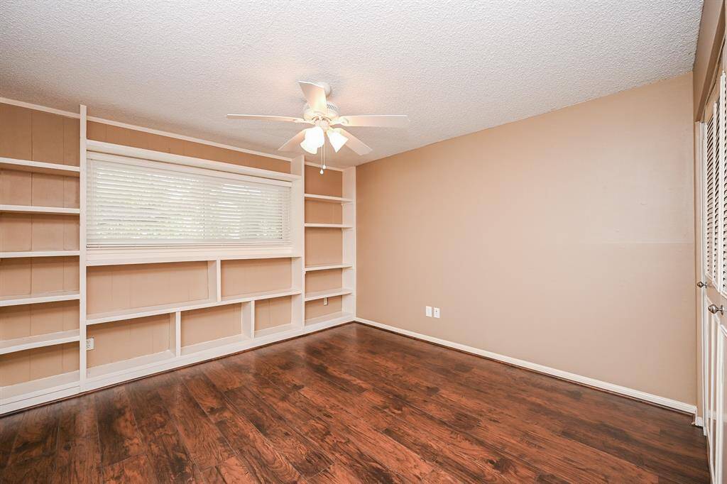 Bright room with dark wood flooring, built-in shelving, a large window with blinds, and a ceiling fan. Ideal for a home office or study.