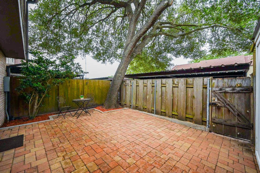 This photo showcases a cozy, fenced backyard with a charming brick patio, shaded by a large tree. There's a  room for a small bistro set for outdoor relaxation and greenery along the edges for a touch of nature.