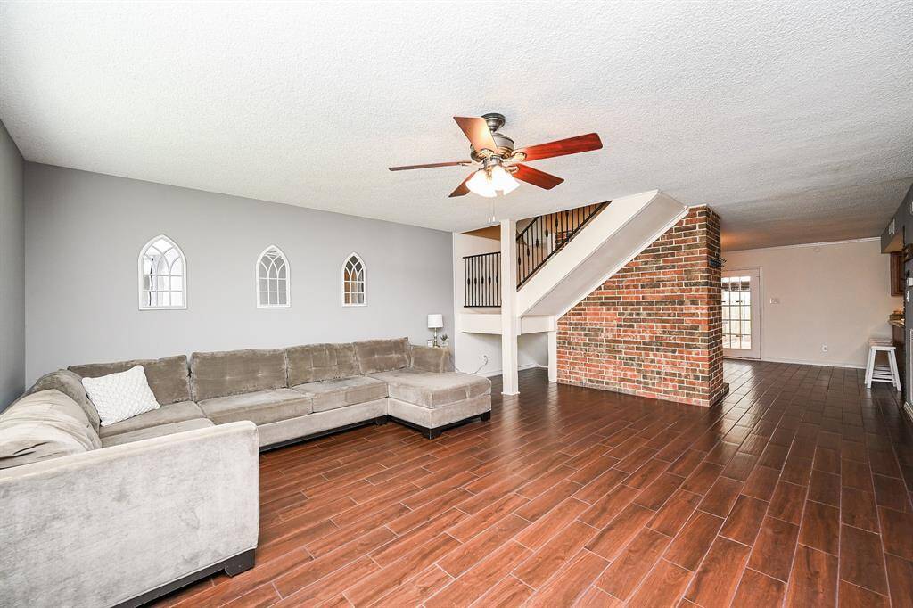 Spacious living area with wood-look tile flooring, neutral gray walls. Features a brick accent wall, ceiling fan, and a staircase leading to the upper level. The open layout flows into a dining area with natural light from French doors.