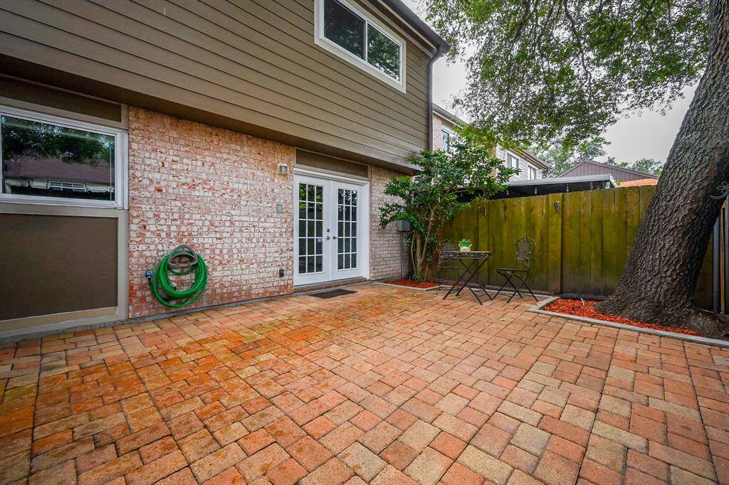 This photo showcases a spacious, paved backyard patio with brick flooring, a charming outdoor seating area, and a large tree providing shade. The space is enclosed by a wooden fence and features French doors leading into the house.