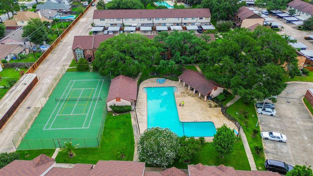 This photo shows a residential complex featuring a large swimming pool with a surrounding patio area, a tennis court, and ample greenery. The area is lined with townhouses and has nearby parking, creating a welcoming community environment.
