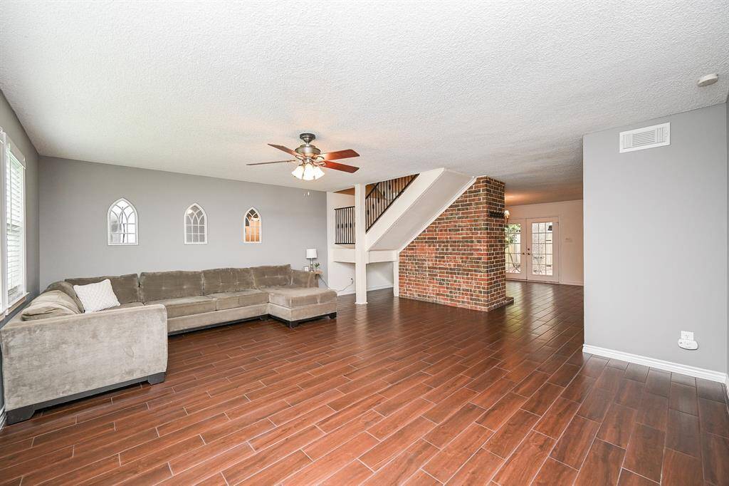 This spacious living room features wood-style flooring, a cozy brick accent wall, and a ceiling fan. It has ample natural light from large windows, a stylish staircase, and an open layout perfect for entertaining.