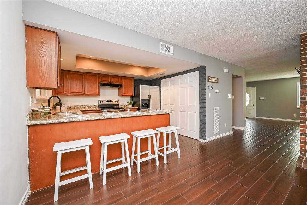 This photo showcases a spacious open-concept kitchen and living area. The kitchen features granite countertops, modern appliances, and a breakfast bar with four stools. The flooring is a rich wood tone, complementing the warm cabinetry, and there's a view into an adjoining bright living space.