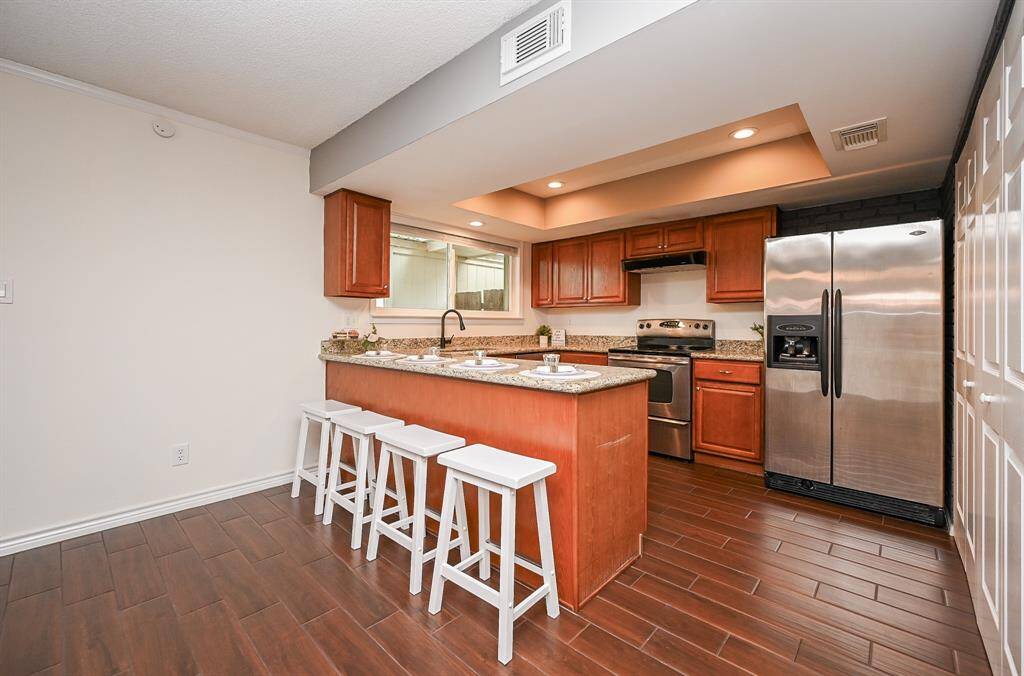 This photo showcases a modern kitchen featuring a central island with granite countertops and four bar stools. It includes stainless steel appliances and warm wooden cabinets, complemented by recessed lighting and wood-look tile flooring.