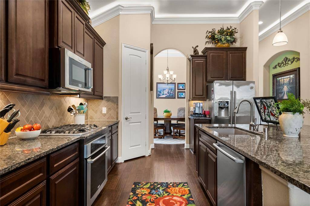 This kitchen features rich dark wood cabinetry, granite countertops, and stainless steel appliances, including a double oven and a microwave. The space opens to a dining area through an arched doorway, offering an elegant chandelier.
