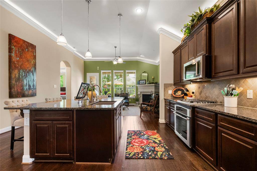 This photo showcases a spacious open-concept kitchen and living area. The kitchen features dark wood cabinetry, granite countertops, and stainless steel appliances, while a large island provides additional seating. The living area is bright with natural light from multiple windows and leads to a cozy fireplace. High ceilings and modern lighting add to the elegant ambiance.