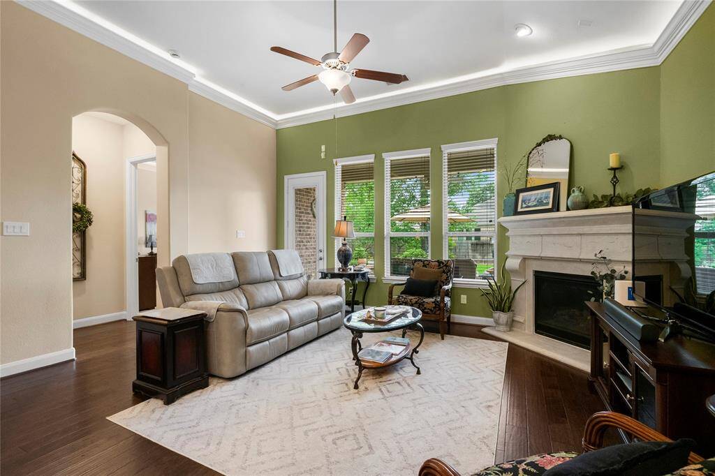 This cozy living room features a vaulted ceiling with crown molding, a ceiling fan, and large windows providing ample natural light. The space includes a comfortable seating area, a stylish fireplace, and a view of the lush backyard. Warm wood flooring and neutral walls complement the inviting atmosphere.