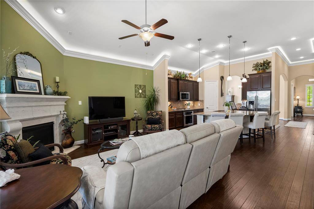 This inviting open-concept living area features a cozy fireplace, elegant ceiling fan, and recessed lighting. It seamlessly connects to a modern kitchen with dark wood cabinets, stainless steel appliances, and a spacious island with pendant lighting. The space is accented with warm wood flooring and tasteful decor.