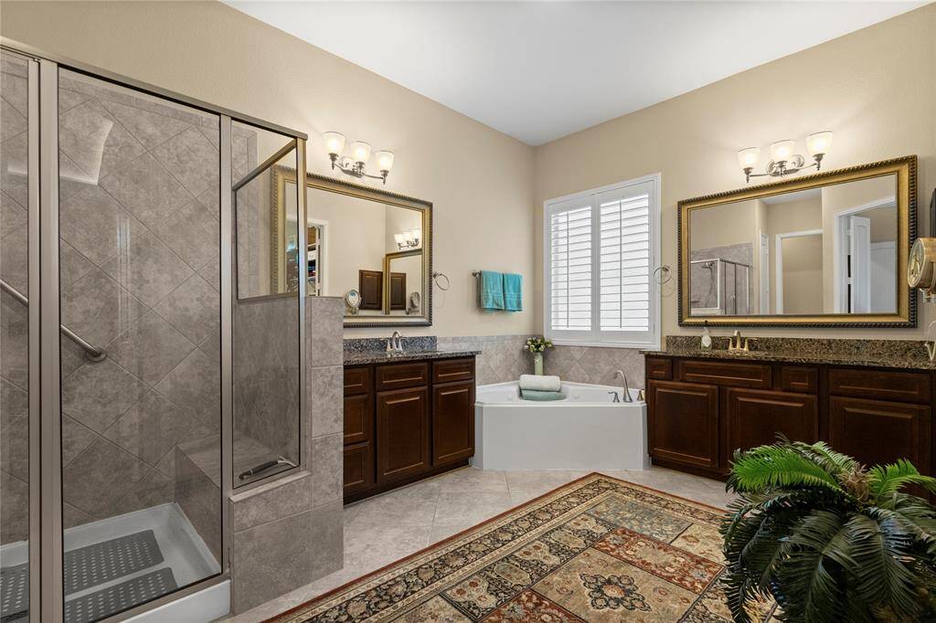 Spacious, elegant bathroom featuring a large glass-enclosed shower, double vanity with granite countertops, and a central soaking tub. Warm lighting, rich wood cabinetry, and a decorative rug add a touch of luxury. Natural light pours in through the window with plantation shutters.