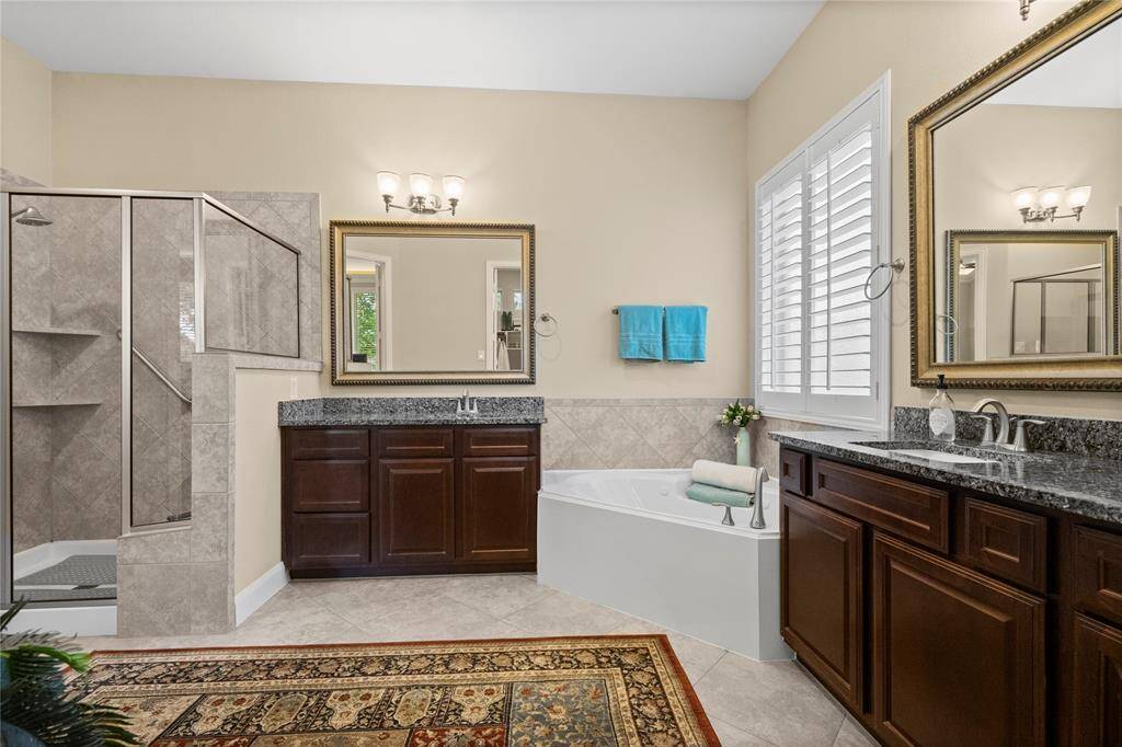This spacious bathroom features a walk-in shower, a soaking tub, and dual vanities with granite countertops. The warm wood cabinetry and elegant mirrors enhance the luxurious feel, complemented by tile flooring and soft lighting.
