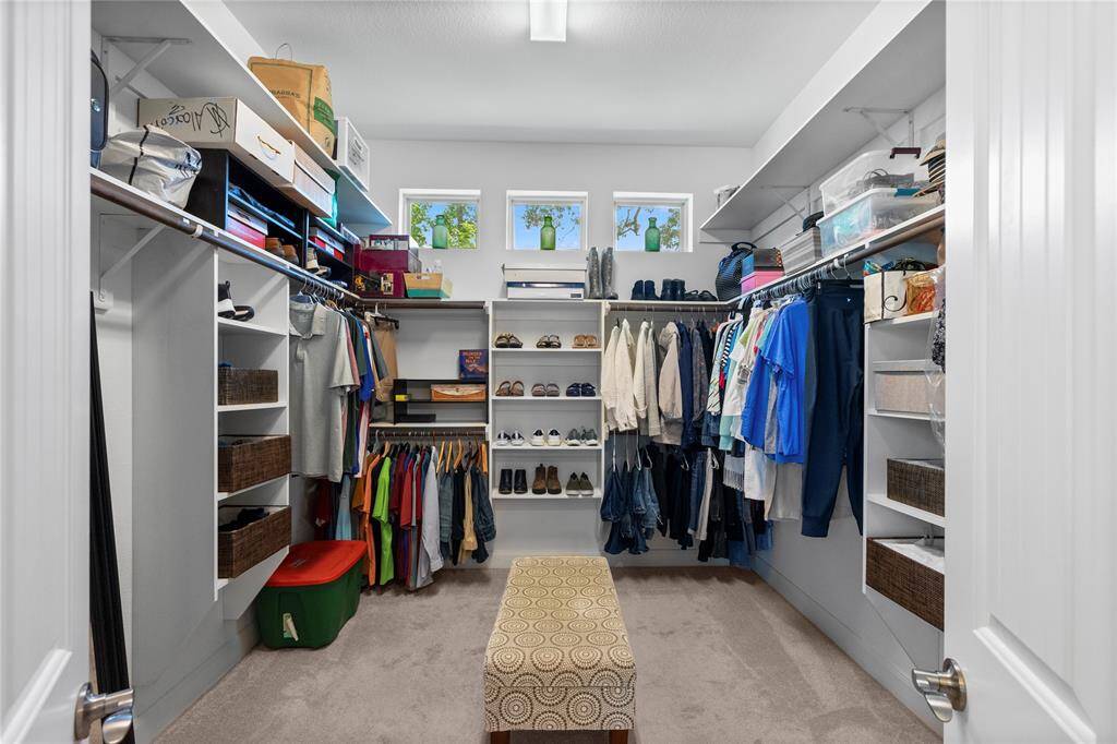 This spacious walk-in closet features ample shelving and hanging space, a central bench, and natural light from three small windows, offering a well-organized and bright storage solution.