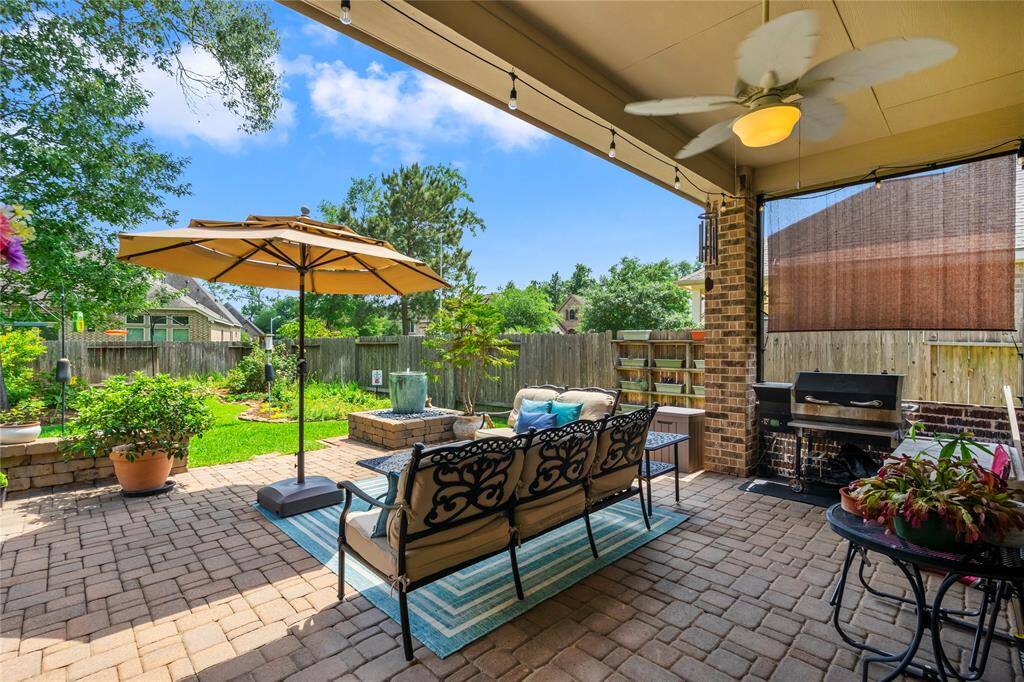 This inviting backyard features a covered patio with a ceiling fan, comfortable seating, and a shade for the grill. It opens to a beautifully landscaped garden with a large umbrella and lush greenery, perfect for relaxing or entertaining.
