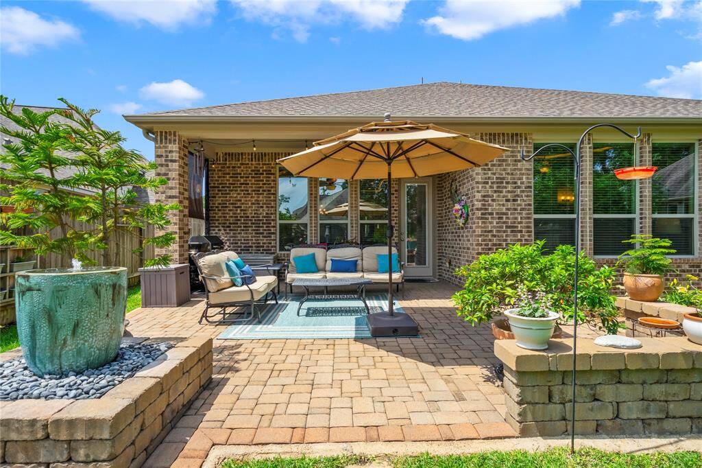 This photo showcases a charming backyard patio with a brick-paved area, cozy seating under a large umbrella, and lush plants. It features a water fountain and offers a relaxing outdoor space, perfect for entertaining or unwinding.