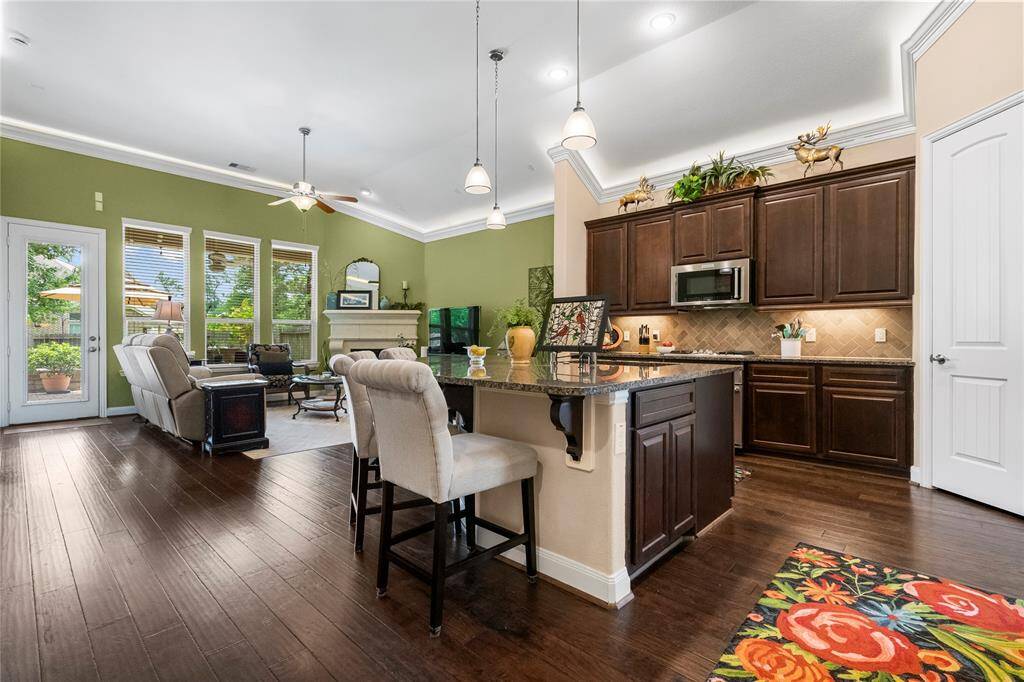 This inviting open-concept living space features a kitchen with dark wood cabinets, granite countertops, and a breakfast bar. The adjacent living area has large windows, abundant natural light, and a cozy seating arrangement. The hardwood floors and green accent wall add warmth and character, while the door leads to an outdoor patio.