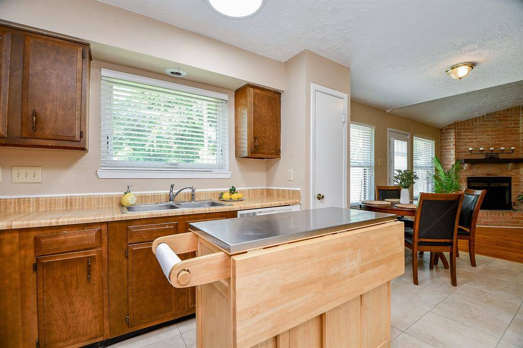 This bright kitchen features wooden cabinetry, a mobile island with a stainless steel top, and a large window for natural light. Adjacent is a cozy dining area with a fireplace, creating a warm, inviting space for meals and gatherings.