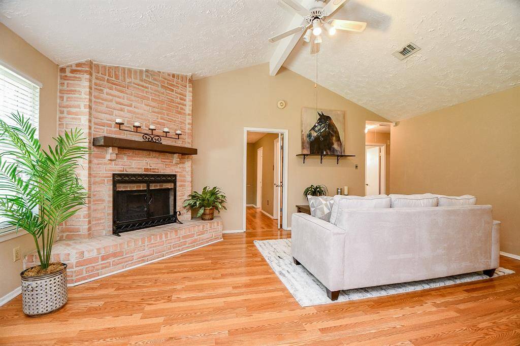 Inviting living room view from kitchen/dining combo features a vaulted ceiling with a fan, a cozy brick fireplace, warm hardwood floors. Natural light streams through the windows.
