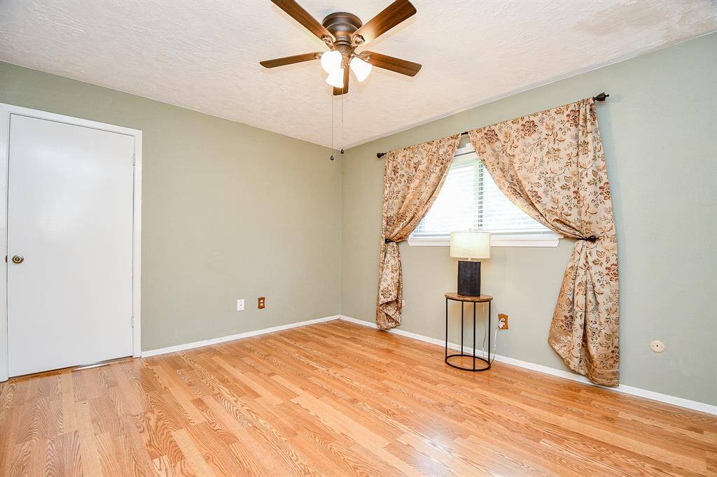 Primary bedroom features hardwood floors, a ceiling fan, and a soft green wall with a window adorned with patterned drapes.  Door leads into living room area offering an inviting private atmosphere.