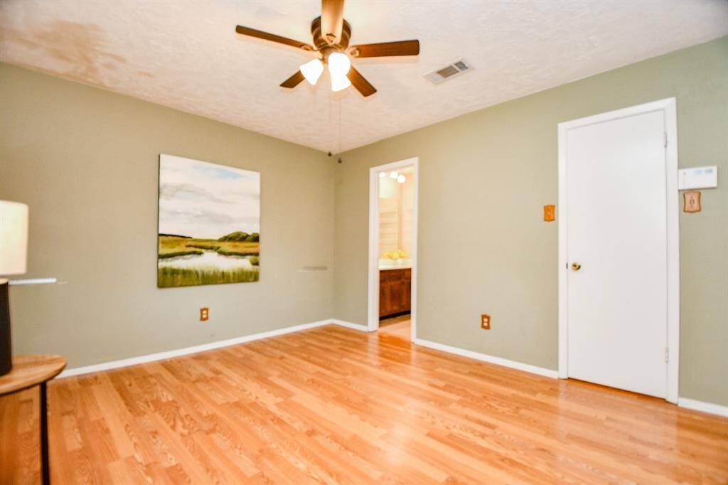 This primary bedroom features warm wooden flooring, a ceiling fan, and soft green walls with a nature-themed painting. It has a doorway leading to a bathroom area and a closed white door leading into a 5X5 walk-in closet.