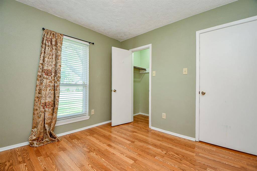 2nd bedroom features hardwood flooring, a soft green wall, a window with floral curtains, and a walk-in closet. It's bright and cozy, perfect for a bedroom!