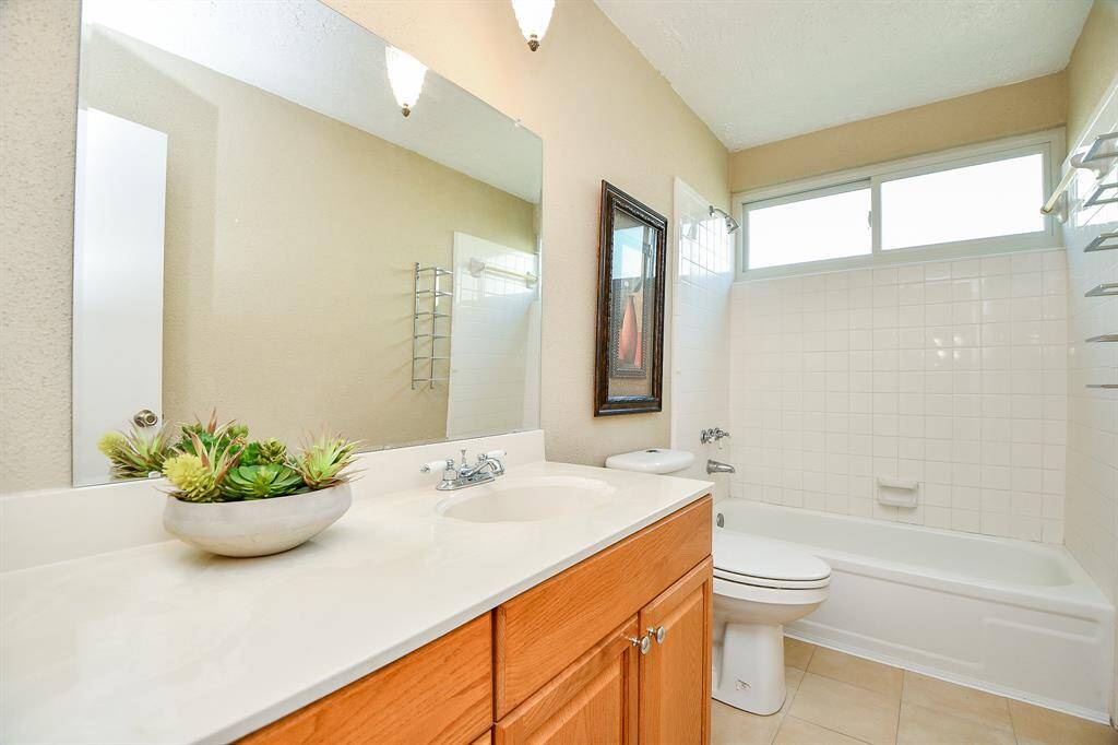 Bright bathroom featuring a large mirror, modern vanity with wooden cabinets, and a white tile bathtub. Natural light pours in through a high window, enhancing the clean, fresh ambiance.