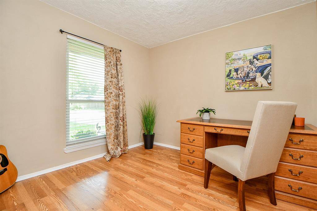 Home office space (3rd bedroom) with warm wood flooring, a large window with floral curtains, and a wooden desk. The room includes a comfortable chair, wall art, and a touch of greenery, creating an inviting atmosphere for work or study.