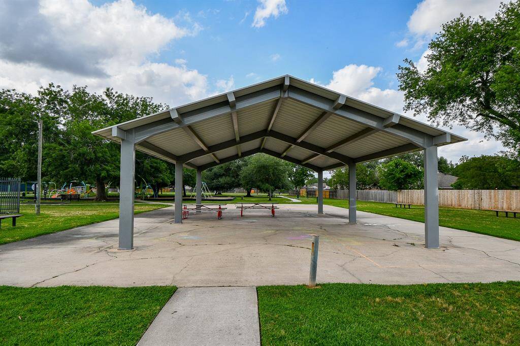 Large, covered pavilion in a park setting, ideal for gatherings. It's surrounded by green lawns and mature trees, with a playground visible in the background, creating a perfect community space for outdoor activities.