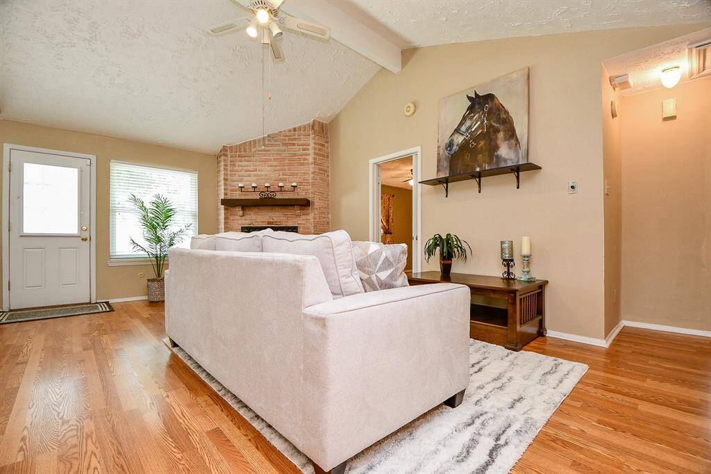 Welcome home to living room features high ceilings, a cozy brick fireplace, and hardwood floors. Natural light streams in through a large window, complementing the neutral-toned walls.A ceiling fan and tasteful decor add to the comfort and style.