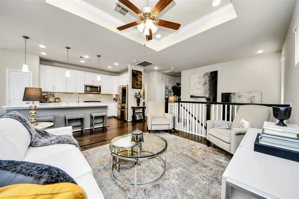 This photo showcases a bright, open-concept living area with elegant hardwood floors and neutral walls. The space features a stairway with dark wood and white railings, leading to a cozy seating area with large windows and abundant natural light. A ceiling fan and modern light fixtures add style, while decorative elements enhance warmth and charm.