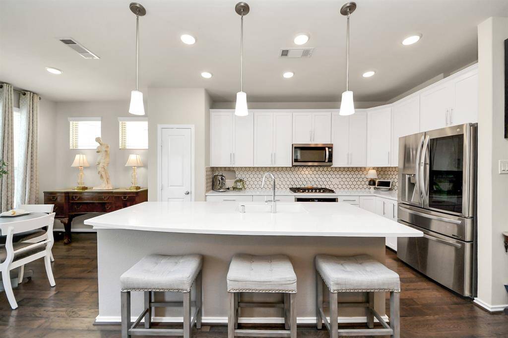 This modern kitchen features a spacious white island with seating, stainless steel appliances, and ample cabinetry. The open layout is enhanced by pendant lighting and a stylish backsplash, creating a bright and inviting space.
