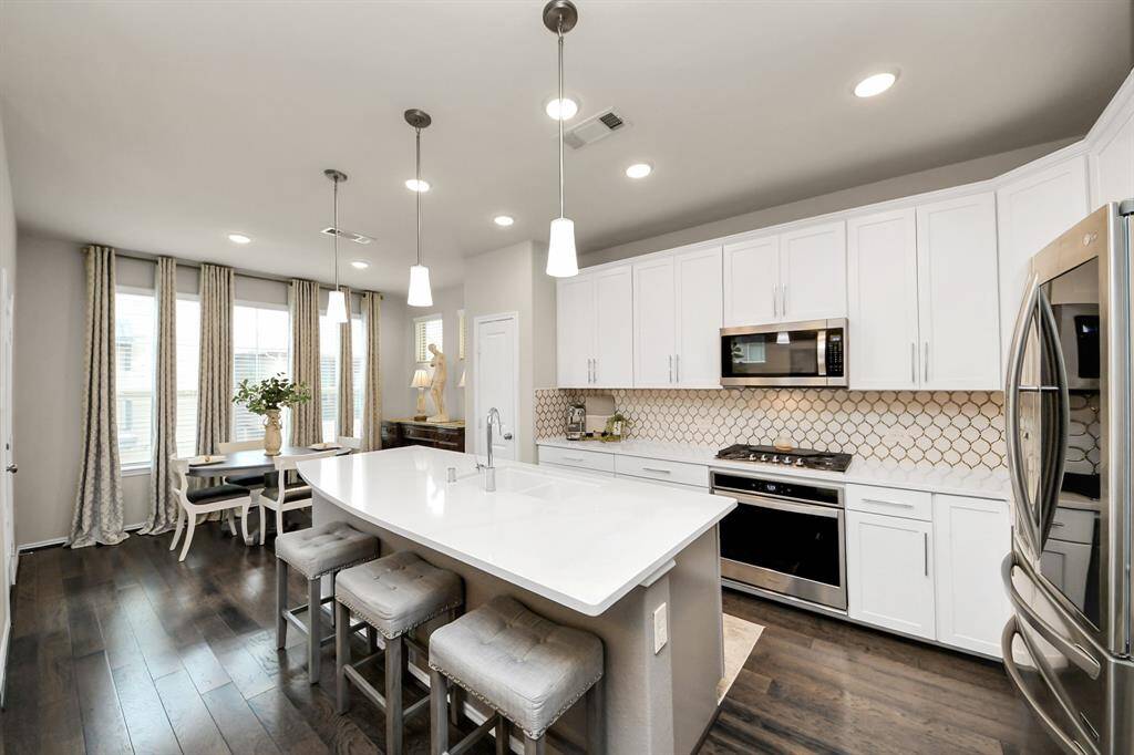 This modern kitchen features sleek white cabinetry, a large island with seating, and stainless steel appliances. It opens into a bright dining area with large windows and elegant drapes, creating a welcoming space for entertaining.