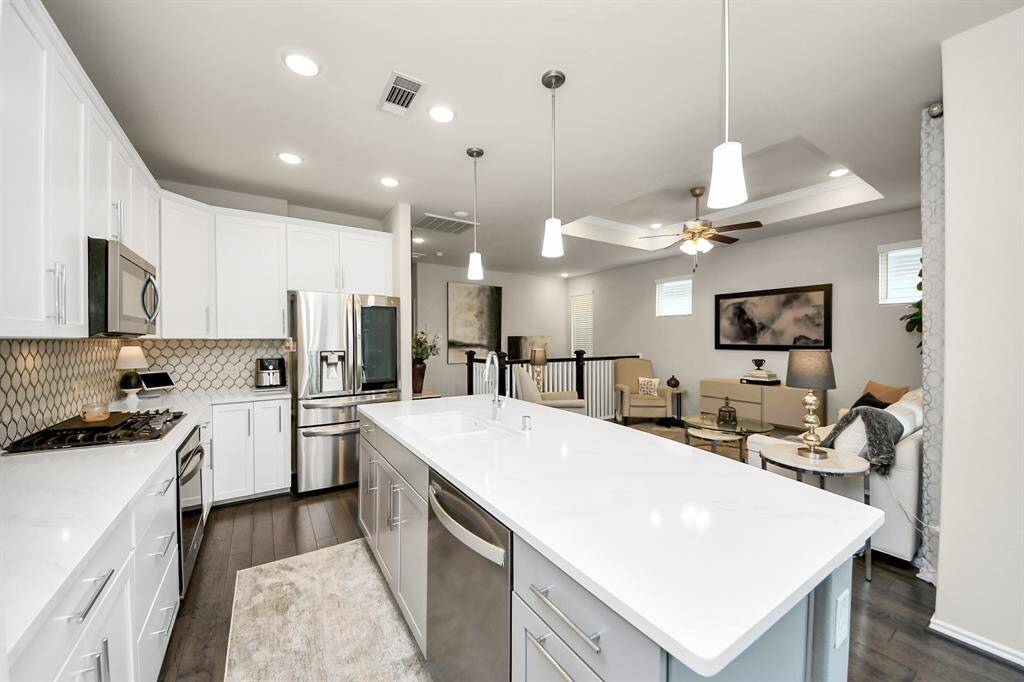 This photo showcases a modern, open-concept kitchen and living area. The kitchen features sleek white cabinetry, stainless steel appliances, and an island with pendant lighting. The living space is cozy, with neutral decor and a ceiling fan, creating a welcoming ambiance.