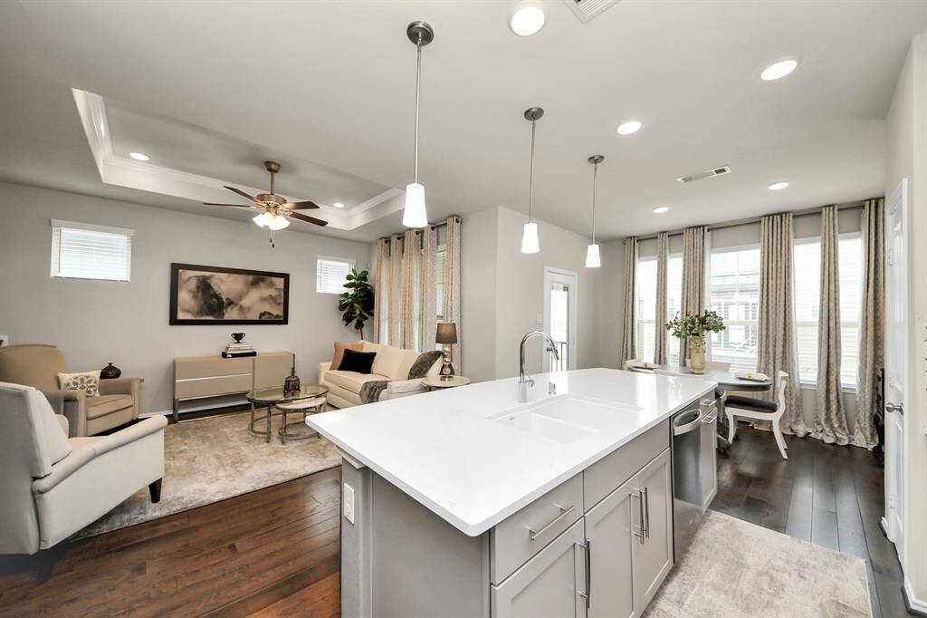 This photo showcases a modern, open-concept living area with a bright kitchen featuring a large island and pendant lighting. The living room includes a cozy seating arrangement, tray ceiling with a fan, and elegant decor. Large windows allow plenty of natural light, complemented by soft, neutral tones throughout.