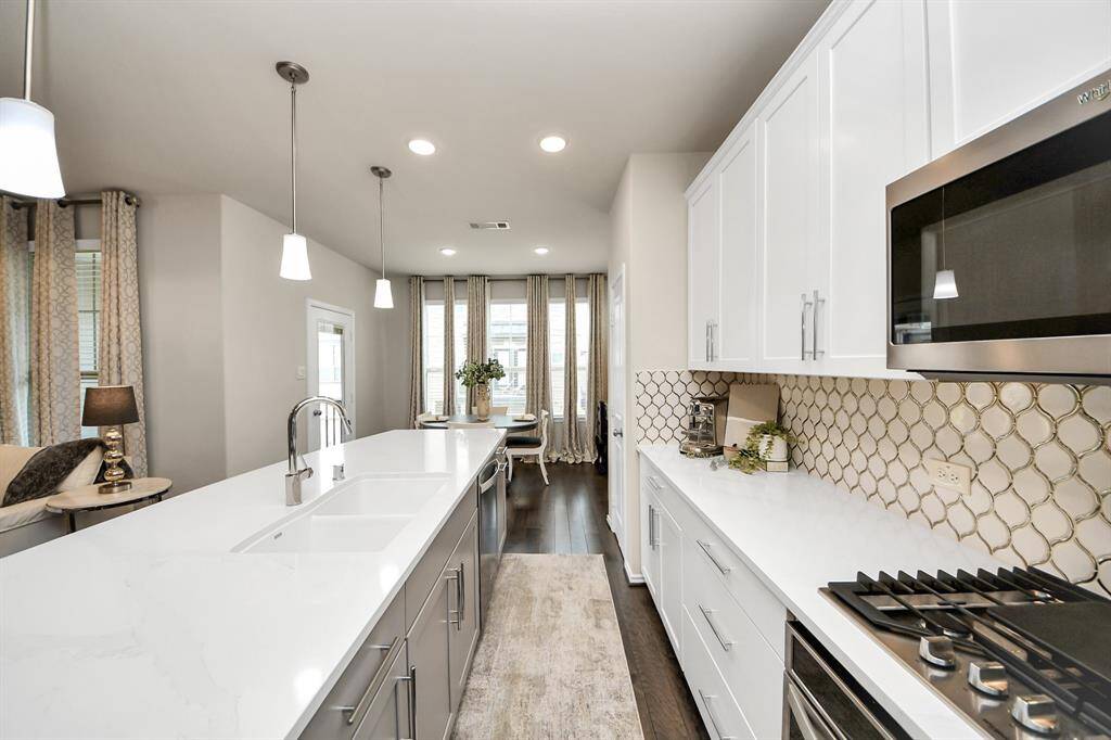 This modern kitchen features a sleek design with white cabinetry, a stylish geometric backsplash, and stainless steel appliances. An expansive island with a sink and pendant lighting provides ample workspace, while the open layout flows seamlessly into a bright dining area.