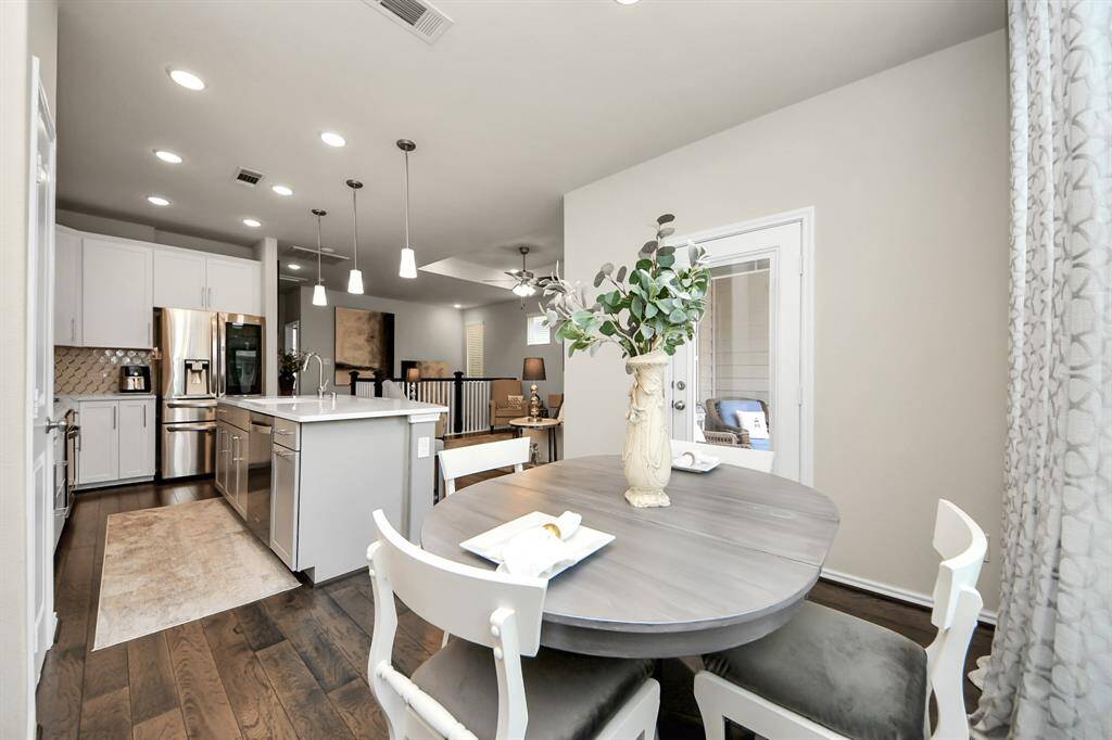 This photo showcases a modern, open-concept kitchen and dining area. It features sleek white cabinetry, stainless steel appliances, and a spacious island with pendant lighting. The dining space includes a round table with stylish chairs, and there's ample natural light from a nearby window with elegant curtains. The dark hardwood floors add warmth to the bright, contemporary design.