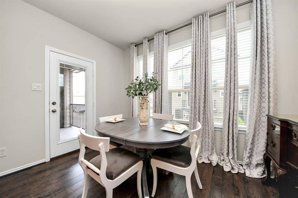 This dining area features a cozy round table with elegant chairs, set against large windows draped with patterned curtains, providing ample natural light. A door leads to a small balcony, enhancing the room's charm.
