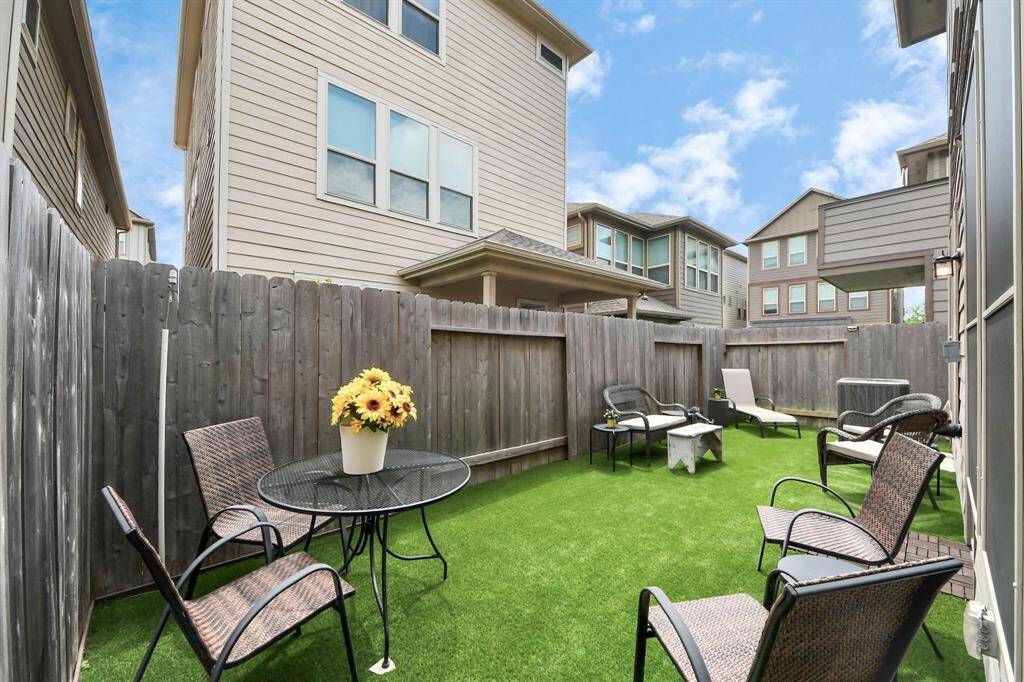 Cozy backyard with artificial turf, surrounded by a wooden fence. Features outdoor seating and a small table, ideal for relaxation or entertaining.