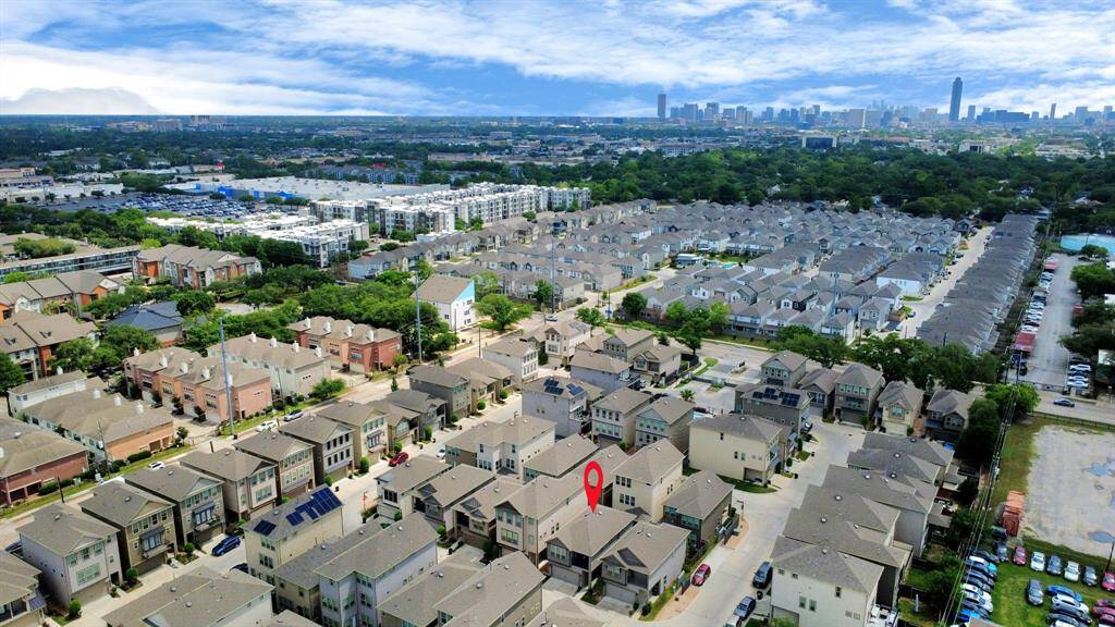 This aerial photo showcases a suburban neighborhood with modern homes, marked by a red pin for easy identification. The area is densely packed with residential buildings, surrounded by greenery and set against a backdrop of a distant city skyline, indicating proximity to urban amenities.